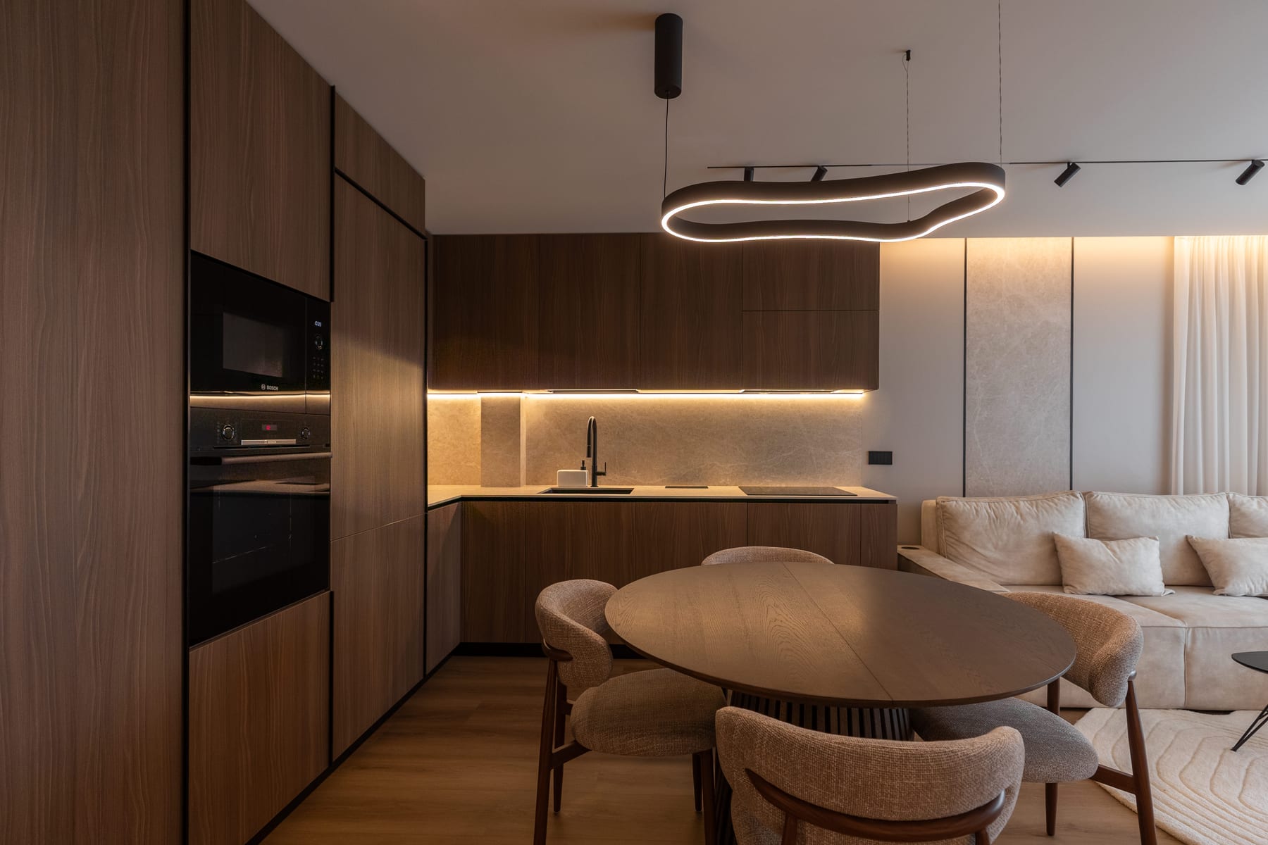 Open-plan kitchen and living area at dusk: full-height walnut cabinetry with an integrated oven, a stone splashback lit by a concealed strip, an oval dining table under a sculptural ring pendant, and a pale sofa beyond.
